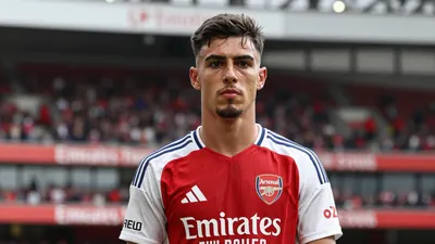 A determined 28-year-old Arsenal striker, wearing the red and navy home kit with the Arsenal crest on his chest, looks...