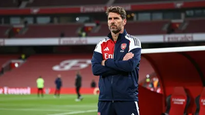 A medium shot of a rugged, 35-year-old Arsenal coach with a stern expression, standing in the Emirates Stadium tunnel...