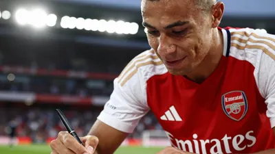A close-up shot of an anonymous Arsenal player in a red jersey with white sleeves, holding a pen and smiling as they sign...
