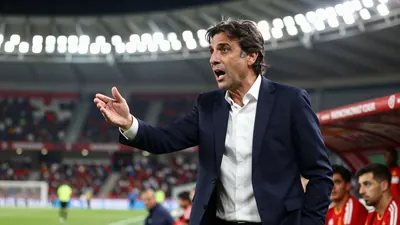 A mid-30s Spanish football manager in a dark blue suit and white shirt, standing on the sidelines during a match...