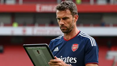 A medium shot of a rugged, 35-year-old, first-team coach, with a strong jawline and receding brown hair, standing in...