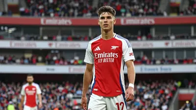 A young Arsenal midfielder in a red and white jersey, standing confidently on the pitch, looking focused with the...