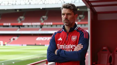 A medium shot of a rugged, 35-year-old, 5'10" Arsenal coach, with a stern expression, standing in the Emirates Stadium...