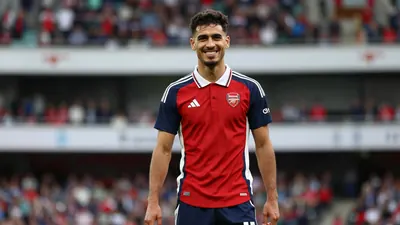 Mid-20s athletic forward, dark hair, confident grin, waist-up in Arsenal home kit - red shirt with navy sleeves, crisp...